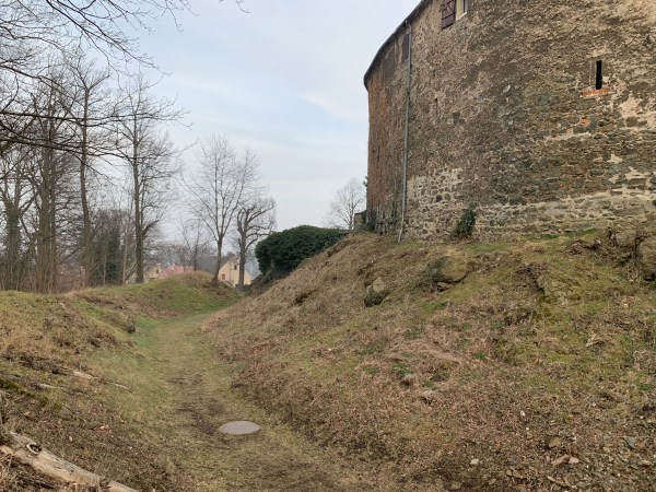 Příkop, vlevo stromy, vpravo hradní zeď.
