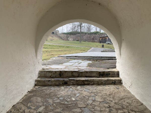 Průchod s bílou zdí na kamenné podlaze, vedoucí ke dvěma kamenným schodištím a vnitřnímu nádvoří.