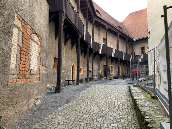 Dlážděná cesta, mírně vzhůru, obklopená zdmi domů, mírná pravotočivá zatáčka, vlevo rytířské dřevěné balkony, vpravo oplocení budovy.