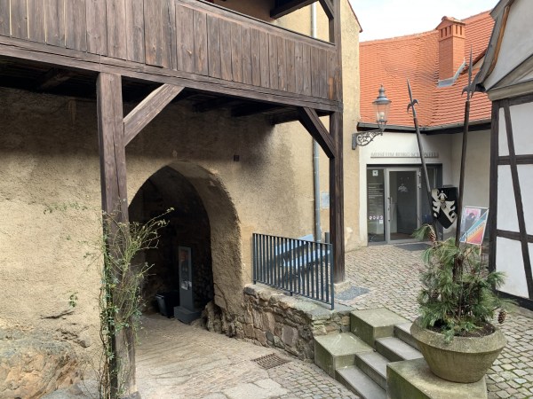 Klikaté zdi domu. Vlevo cesta kulatým obloukem, nad ní dřevěný balkon. Vpravo 4 schody na vyšší úroveň s cestou k proskleným dveřím, nad nimi červená střecha.