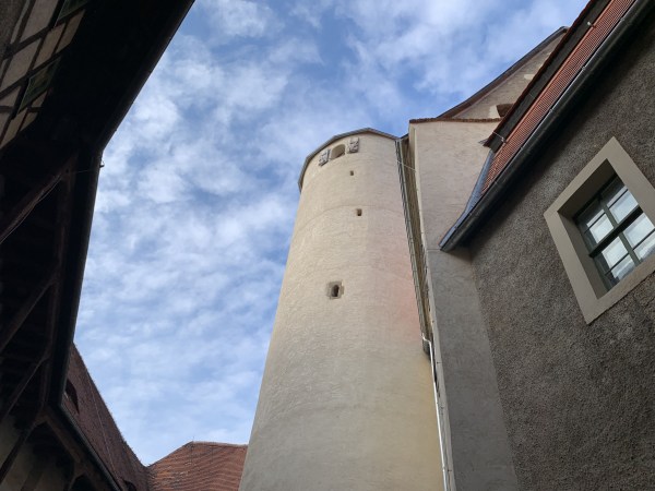 Béžová omítnutá hradní věž s téměř žádnými otvory, pohled zespodu, obklopená budovami za mírně zamračeného počasí