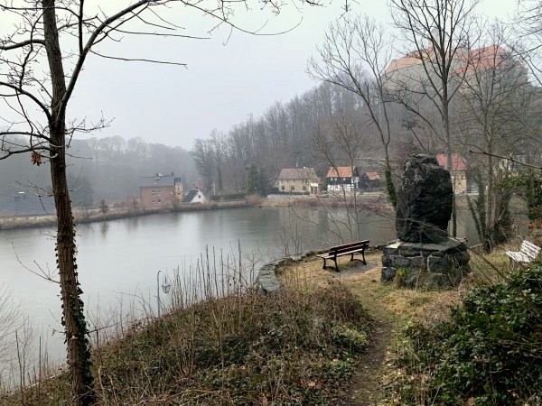 Stezka u rybníka s lavičkou za mlhavého počasí. Za lavičkou se nachází balvan. Za rybníkem stromy a domy. Hrad na kopci vpravo.