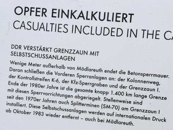 Tabulka s nápisem: OPFER EINKALKULIERT - NDR posiluje hraniční plot automatickými střeleckými systémy. Vysvětlení k zábranám.