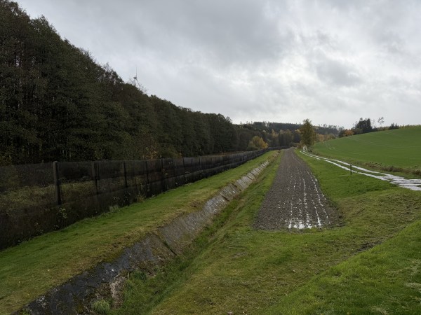 Za polem hraniční pás s příkopem a plotem, za ním stromy