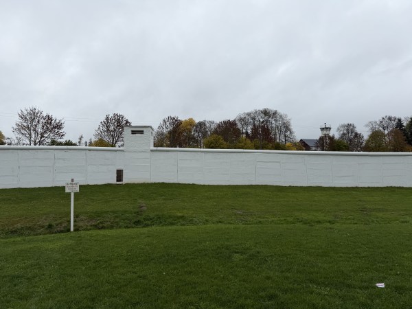 Za zamračeného počasí bílá zeď za loukou a příkopem. Na louce je umístěna cedule. V pozadí stromy a hraniční věž.