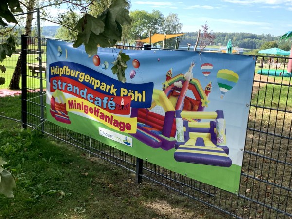 Bunter Banner mit blauem Hintergrund am Zaun mit geschwungener Aufschrift Hüpfburgenpark Strandcafe Minigolfanlage. Neben der Schrift auf dem Banner sind Hüpfburgen abgebildet. Hinter dem Zaun deutet sich ein Freizeit-Areal an.