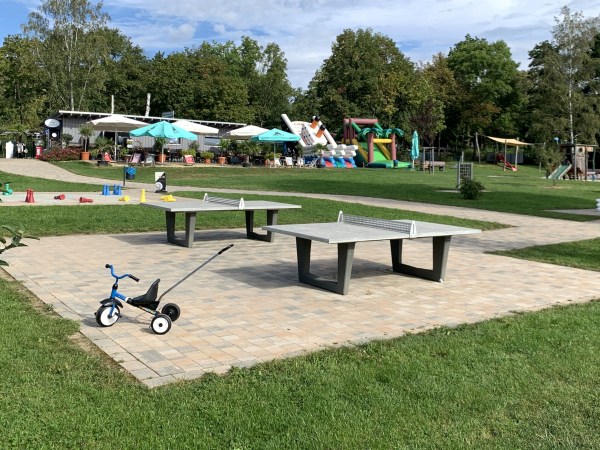 Zwei Tischtennis-Platten auf einer Wiese. Vorne am Steinrand zur Wiese steht ein Dreirad mit Schiebestange. Im Hintergrund weitere Spielgeräte, Hüfburgen, Sonnenschirme und Wald