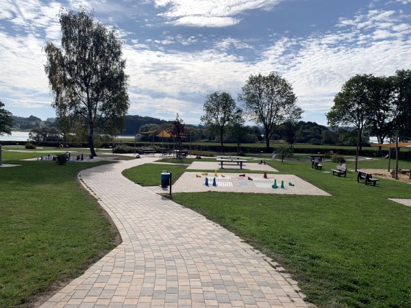 Geschlängelter Weg durch Park mit fest installietrten Outdoor-Spielen, Bänken und vereinzelt Bäume.